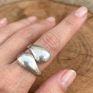 Op de afbeelding: Een zilverkleurige ring met twee gladde, druppelvormige elementen die elkaar licht overlappen. Het metaal heeft een gepolijst, reflecterend oppervlak met een gebogen, organisch ontwerp dat om de vinger wikkelt voor een vloeiende uitstraling.
