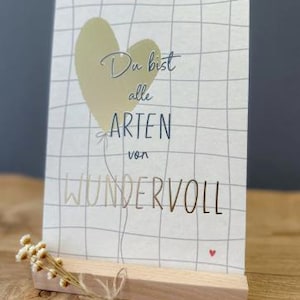 Può includere: Un biglietto d'auguri rettangolare con sfondo bianco, un motivo a griglia grigio, un palloncino a forma di cuore giallo pallido e il testo in tedesco Du bist alle ARTEN von WUNDERVOLL. Il biglietto poggia su un supporto in legno chiaro.
