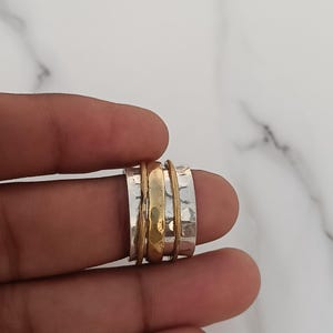 May include: A wide spinner ring featuring a central polished gold-tone band flanked by two outer silver-tone bands. The silver-tone bands have a textured, hammered finish, while the gold-tone band is smooth and rounded in the center.