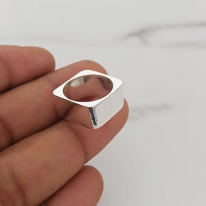 Puede incluir: Un anillo de tono plateado pulido que presenta una banda gruesa de forma cuadrada con una abertura circular en el centro. La superficie metálica es lisa y reflectante, mostrando un diseño geométrico con esquinas redondeadas y una construcción sólida.