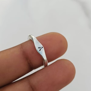 Può includere: Un anello color argento con una superficie frontale liscia e leggermente appiattita, caratterizzato dalla lettera A nera stampata al centro. La fascia presenta una finitura lucida e riflettente e una forma sottilmente ondulata che si restringe.