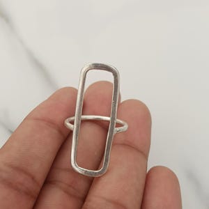 Könnte beinhalten: Ein silberfarbener Ring mit einem großen, vertikal ausgerichteten rechteckigen Rahmendesign. Der Ring hat eine glatte, metallische Oberfläche und ein dünnes Band, das an den Seiten der länglichen Rechteckform ansetzt und einen offenen Look erzeugt.