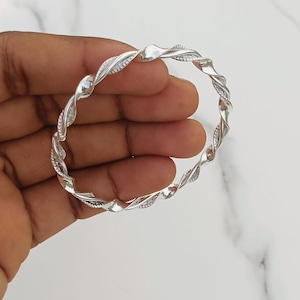 May include: A silver-toned bangle bracelet featuring a continuous twisted rope design. The metal surface alternates between smooth, reflective sections and textured, patterned segments that catch the light, creating a dynamic visual effect.