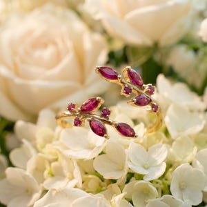 May include: A gold-toned open ring featuring two branch-like segments adorned with marquise-cut and round-cut deep pinkish-red gemstones. The design mimics a natural vine with leaves, with the stones set along the metallic branches.