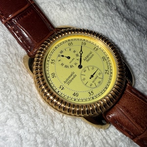 Op de afbeelding: Een Admiral Nelson automatisch horloge met een gele wijzerplaat, goudkleurige geribbelde rand en bruine lederen band. De wijzerplaat bevat een sub-wijzerplaat voor uren met Romeinse cijfers, een sub-wijzerplaat voor seconden en de tekst Automatic 20 Jewels.