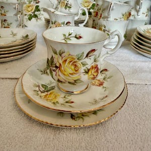 May include: A white porcelain teacup and saucer set featuring a yellow rose pattern with green leaves and gold trim. The teacup has a scalloped rim and a curved handle, while the matching saucer has a scalloped edge with a gold-painted border.