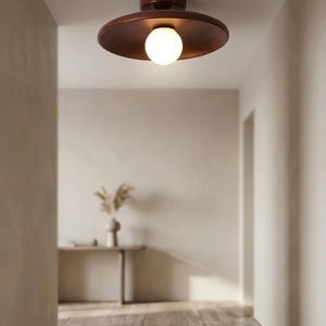 Puede incluir: Una lámpara de techo de montaje empotrado con una base circular de madera marrón oscuro y una pantalla ancha y plana en forma de disco de madera marrón oscuro. Una bombilla esférica de cristal esmerilado blanco está centrada bajo el disco.