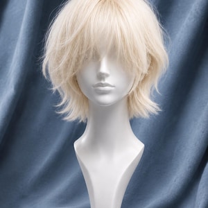 Könnte beinhalten: Eine kurze, gestufte, platinblonde Perücke mit fransigem Pony und strukturierten Spitzen, präsentiert auf einem glatten weißen Schaufensterpuppenkopf. Das Haar hat einen hellen, aschblonden Farbton mit weichem, gefiedertem Aussehen.