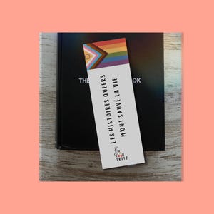 Peut inclure: Un marque-page rectangulaire blanc avec un motif de drapeau Progress Pride en haut. Le texte "LES HISTOIRES QUEERS M'ONT SAUVÉ LA VIE" est imprimé en noir, suivi d'un petit logo avec le texte "BW LOU TRITZ" et deux cerises rouges.