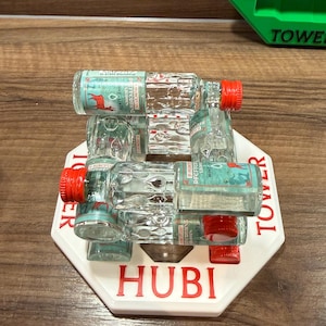 Könnte beinhalten: Ein weißer achteckiger Sockel mit dem roten Schriftzug HUBI TOWER trägt einen Stapel kleiner, klarer Glasflaschen mit roten Verschlüssen. Jede Flasche hat ein Etikett mit einem roten Hirsch und dem Text ST. HUBERTUS PFEFFERMINZ TROPFEN MINT LIKOR.