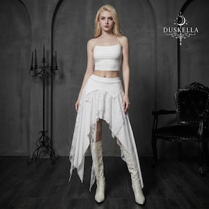 Op de afbeelding: Een witte tweedelige outfit bestaande uit een mouwloze crop top met dunne bandjes en een asymmetrische rok met kanten panelen. De rok heeft een aansluitende tailleband en gelaagde stof, gecombineerd met witte kniehoge laarzen.