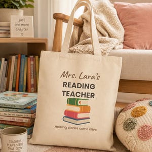 May include: A beige canvas tote bag featuring the text "Mrs. Lara's READING TEACHER" and "Helping stories come alive" in dark brown and black lettering, with a graphic illustration of a stack of four colorful books in green, orange, red, and blue.