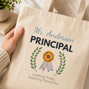 Op de afbeelding: Een beige canvas draagtas met de tekst Mr. Anderson PRINCIPAL in blauw en zwart, een afbeelding van een oranje onderscheidingslint tussen twee groene lauriertakken en de tekst Leading Today, Inspiring Tomorrow eronder.