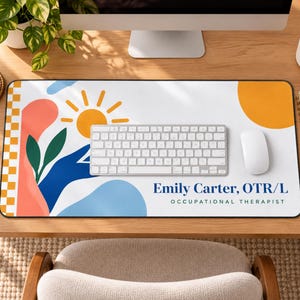 Op de afbeelding: Een rechthoekige bureaumat met een witte achtergrond en abstracte vormen in koraal, blauw en geel, inclusief een zon- en bladgrafiek. De tekst luidt Emily Carter, OTR/L OCCUPATIONAL THERAPIST in donkerblauwe en zwarte letters.