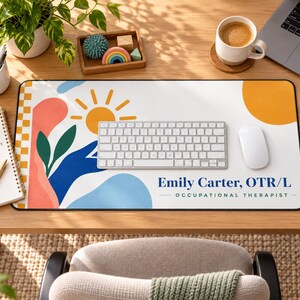 Op de afbeelding: Een rechthoekige bureaumat met een witte achtergrond, een gele zon, abstracte blauwe en koraalkleurige vormen en een geblokte rand. De tekst luidt Emily Carter, OTR/L OCCUPATIONAL THERAPIST. Een zilverkleurig toetsenbord en witte muis liggen op de mat.