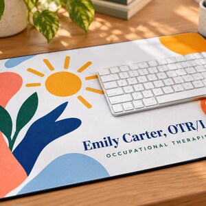 Op de afbeelding: Een rechthoekige bureaumat met een witte achtergrond, een gele zon, groene bladeren en abstracte vormen in perzik, blauw en oranje. De linkerrand heeft een geel-wit geblokt patroon. De tekst luidt Emily Carter, OTR/L OCCUPATIONAL THERAPIST.