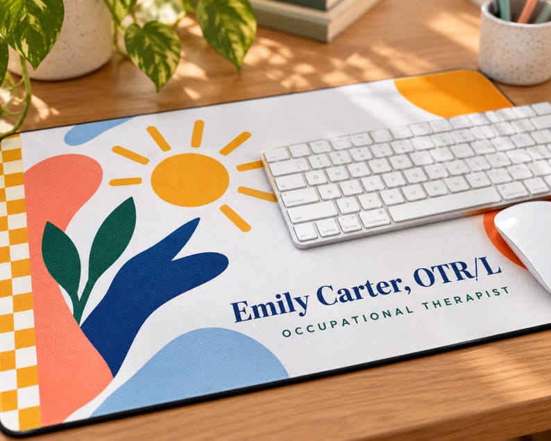 Op de afbeelding: Een rechthoekige bureaumat met een witte achtergrond, een gele zon, groene bladeren en abstracte vormen in perzik, blauw en oranje. De linkerrand heeft een geel-wit geblokt patroon. De tekst luidt Emily Carter, OTR/L OCCUPATIONAL THERAPIST.