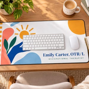 Op de afbeelding: Een rechthoekige bureaumat met een witte achtergrond, een gele zon, een blauwe hand, groene bladeren en oranje vormen. De linkerrand heeft een geel-wit schaakbordpatroon. De tekst luidt Emily Carter, OTR/L OCCUPATIONAL THERAPIST.