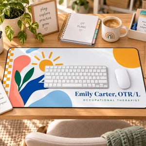 Op de afbeelding: Een rechthoekige bureaumat met een witte achtergrond, abstracte vormen in oranje, roze, blauw en groen, een gele zon en de tekst Emily Carter, OTR/L OCCUPATIONAL THERAPIST in donkerblauw en zwart aan de rechteronderkant.