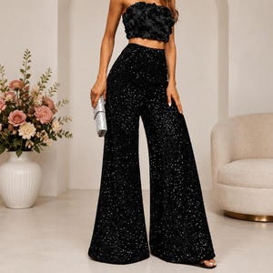 May include: A black strapless crop top featuring a dense arrangement of fabric roses and matching high-waisted wide-leg trousers covered in black sequins. The ensemble is paired with a small, rectangular, silver-toned metallic clutch bag.