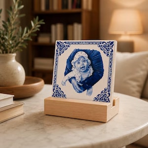 Könnte beinhalten: Eine quadratische Keramikfliese mit einer blau-weißen Illustration eines Babys mit Häubchen, das von Händen gehalten wird. Die Fliese hat einen verzierten blauen Rand und steht aufrecht in einem rechteckigen hellen Holzständer.