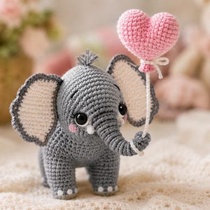 Può includere: Un giocattolo a forma di elefante lavorato all'uncinetto in grigio con grandi orecchie dal centro color crema, occhi di sicurezza neri e guance rosa. L'elefante tiene nella proboscide un piccolo palloncino a cuore rosa chiaro.