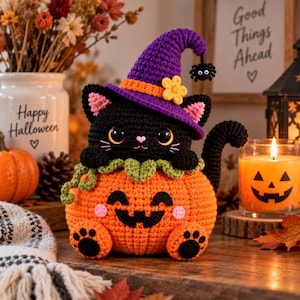 Puede incluir: Un gato negro de ganchillo con un sombrero de bruja morado con una flor amarilla y una pequeña araña, sentado dentro de una calabaza de ganchillo naranja con cara de jack-o'-lantern. El gato tiene ojos grandes, orejas rosas y patas negras.