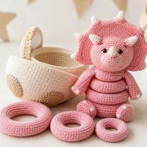 Puede incluir: Un juguete apilable de tricératops rosa tejido a ganchillo con una gran gola y cuernos, acompañado de tres anillos circulares rosas de diferentes tamaños y una cesta tejida de color beige con asa, todo en hilo texturizado.