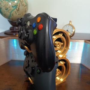 May include: A black video game controller resting on a gold-colored, dual-tiered circular stand. The controller features a black body with multi-colored action buttons in blue, green, yellow, and red. The stand has a metallic gold finish with intricate circular cutouts.