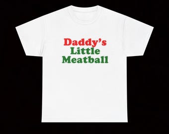 May include: A white short-sleeved t-shirt featuring the text "Daddy's Little Meatball" printed on the front. The word "Daddy's" is in red, while the words "Little Meatball" are in green, all centred in a bold, sans-serif font style.
