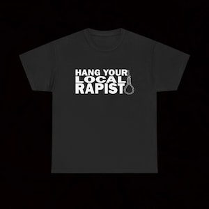 May include: A black short-sleeved t-shirt featuring white text that reads HANG YOUR LOCAL RAPIST, accompanied by a graphic illustration of a hangman's noose positioned to the right of the word RAPIST, centred on the chest area of the garment.