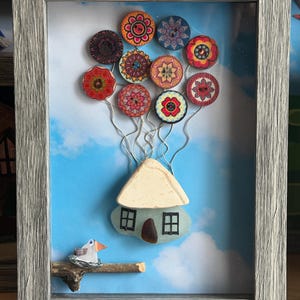 May include: A framed wall art piece featuring a house made of sea glass and ceramic, lifted by colorful patterned buttons attached with silver wire. A small bird sits on a wooden branch below the house against a blue sky background.