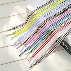Può includere: Una collezione di lacci piatti con motivo a quadretti vichy in vari colori, tra cui marrone, giallo, azzurro, lavanda, rosa, blu scuro, verde, rosso, grigio e nero, ciascuno rifinito con un bordo smerlato di colore bianco.