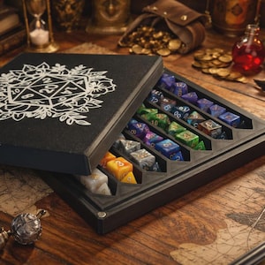Könnte beinhalten: Eine schwarze, rechteckige Würfelaufbewahrungsbox mit einer weißen Grafik aus Blumen und einem 20-seitigen Würfel auf dem Deckel. Die geöffnete Box zeigt mehrere Reihen bunter polyedrischer Würfel in passgenauen Fächern in Blau, Grün und Gelb.