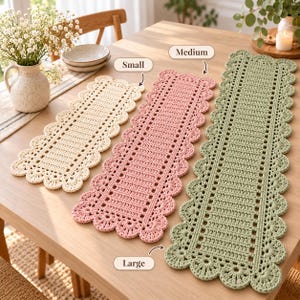 Può includere: Tre runner da tavola rettangolari all'uncinetto in color crema, rosa antico e verde salvia. Ogni runner presenta un motivo centrale a griglia con un bordo smerlato a punto conchiglia. Le etichette indicano le taglie Small, Medium e Large.