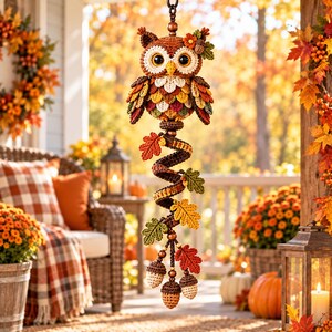 May include: A crocheted owl hanging ornament featuring a brown body with large eyes, a yellow beak, and colorful leaf-shaped wings. Below the owl, a spiral structure is adorned with crocheted autumn leaves and three small acorn-shaped pendants.