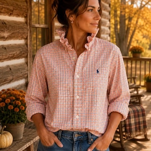 May include: A long-sleeved button-down shirt featuring a small orange, blue, and white plaid pattern, a ruffled collar, and a small dark blue embroidered logo on the left chest, paired with blue denim jeans.