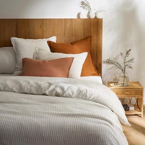 Puede incluir: Una cama con funda nórdica blanca de finas rayas verticales negras, dos almohadas blancas grandes a rayas, dos cojines cuadrados de color óxido y un cojín rectangular de color óxido frente a un cabecero de madera con listones.