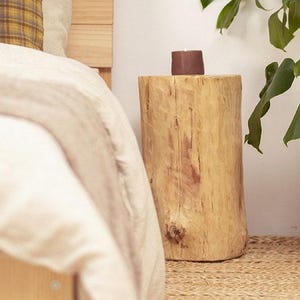 Può includere: Un tavolino cilindrico ricavato da un tronco di legno con finitura naturale marrone chiaro, dotato di una superficie superiore liscia che sostiene una piccola candela marrone scuro. Il tavolo è realizzato in legno massello con venature.