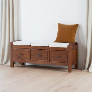 Puede incluir: Un banco de almacenamiento rectangular de madera con tres cajones y tiradores de metal oscuro. El banco tiene tres cojines de asiento blancos en la parte superior y un cojín decorativo cuadrado de color marrón en el lado derecho.