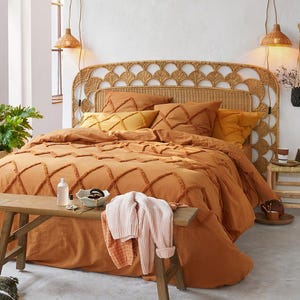May include: A bed with a large arched rattan headboard and a burnt orange tufted duvet cover featuring a diamond pattern. A wooden bench sits at the foot of the bed, holding a light pink sweater, a small white bowl, and a clear glass bottle.