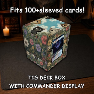 May include: A square TCG deck box with a floral and cat pattern, featuring a clear window displaying a card. The box has text that reads Fits 100+sleeved cards! and TCG DECK BOX WITH COMMANDER DISPLAY. The design shows cats peeking from flowers.