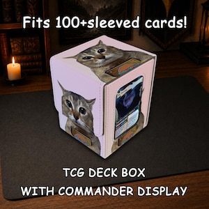 May include: A light pink TCG deck box with a commander display window, featuring a printed cat graphic and the text Fits 100+sleeved cards! TCG DECK BOX WITH COMMANDER DISPLAY. The box has a textured surface and holds a single trading card.