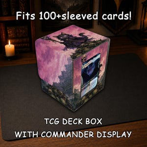 May include: A square TCG deck box with a commander display window, featuring a purple and pink illustration of a cat wizard on a skull-covered landscape. The text reads: Fits 100+sleeved cards! TCG DECK BOX WITH COMMANDER DISPLAY.