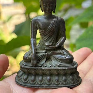 May include: A dark bronze-toned metal figurine of a seated Buddha figure. The figure is depicted in a meditative pose with one hand resting on the lap and the other reaching towards the base. It sits upon a pedestal with a carved lotus petal design.