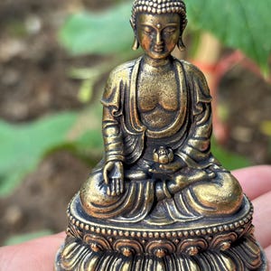 May include: A small, antique-style bronze-colored Buddha figurine in a seated meditation pose. The figure features a textured ushnisha, draped robes, and rests upon a detailed lotus pedestal base with a small lotus flower held in the left hand.