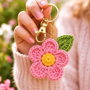 Puede incluir: Un llavero de flor de ganchillo con cinco pétalos rosas, un centro amarillo y una hoja verde. La flor está unida a una anilla y un mosquetón de metal dorado. Aparece el texto Bloom Pattern Creation y Handmade with love, just for you.