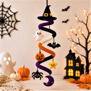 Può includere: Una decorazione a spirale all'uncinetto da appendere, sormontata da un cappello da strega nero. La spirale sfuma dal nero al bianco, all'arancione e al viola. Sono attaccate piccole figure all'uncinetto, tra cui un fantasma bianco, un pipistrello nero, una zucca arancione, una stella gialla, un ragno nero e una mezzaluna viola.