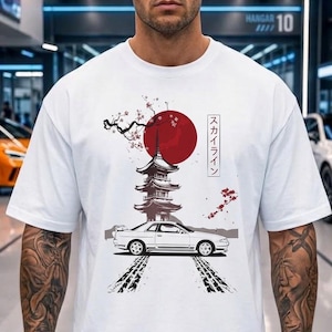 Może przedstawiać: Biały t-shirt z krótkim rękawem z nadrukiem graficznym przedstawiającym biały samochód sportowy, wielopoziomową pagodę, duże czerwone koło i gałązki wiśni. Tekst "スカイライン" jest wydrukowany pionowo w czarnej prostokątnej ramce.