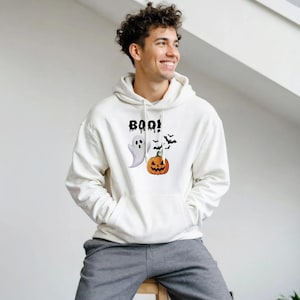 Puede incluir: Una sudadera con capucha de color crema que presenta un estampado gráfico en el pecho. El diseño incluye el texto negro BOO! sobre un fantasma blanco, una calabaza naranja y un pequeño grupo de murciélagos negros volando.